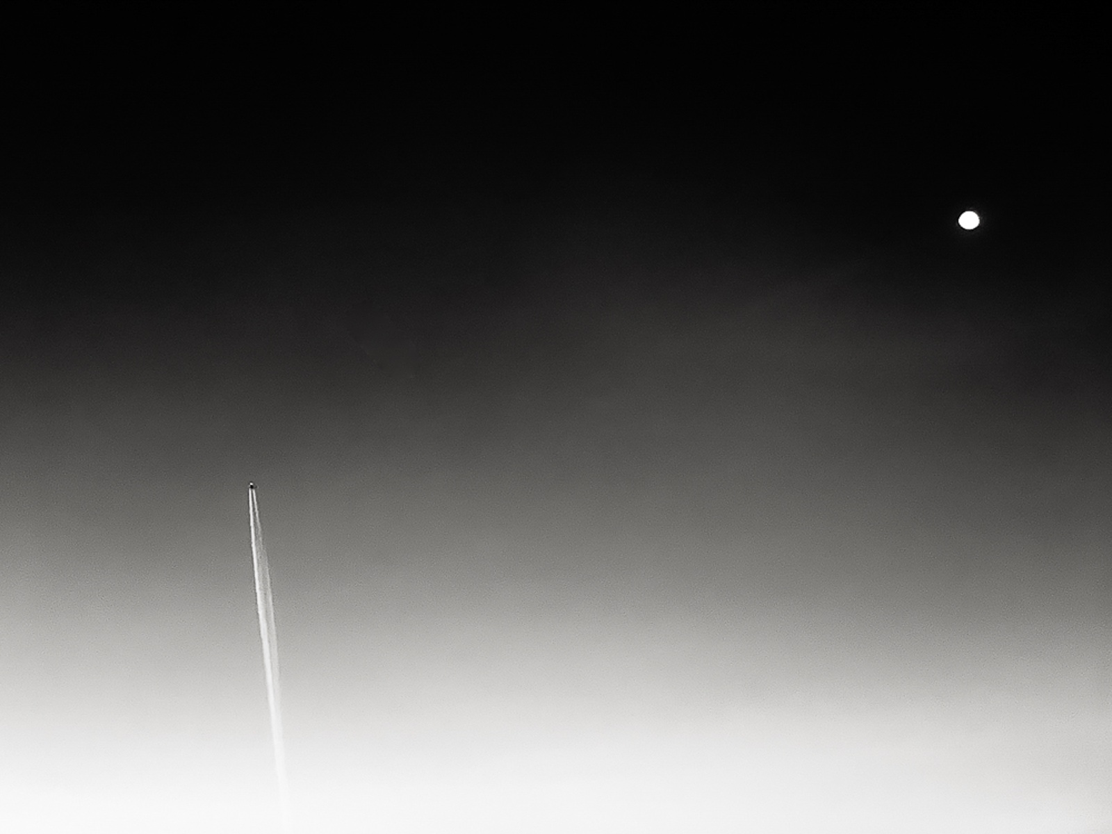 Estela de condensación que sube en diagonal por un cielo degradado de blanco a negro, con la luna en lo alto a la derecha.