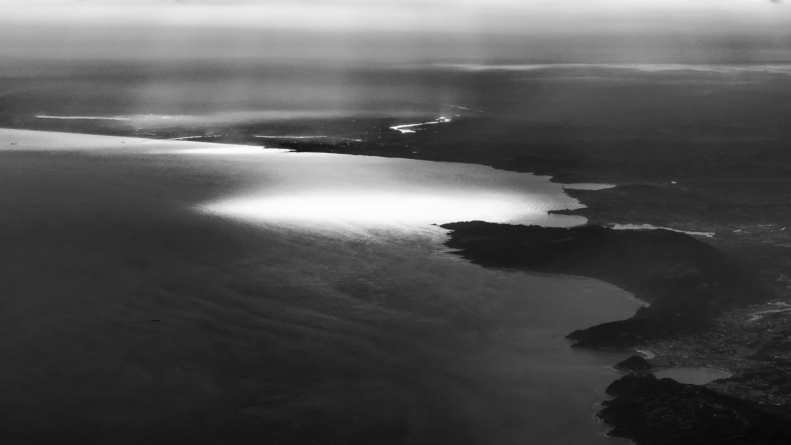 Vista aérea de una costa al contraluz: el sol se refleja en una mancha blanca sobre el mar y la tierra queda en sombra, con una bahía al fondo.