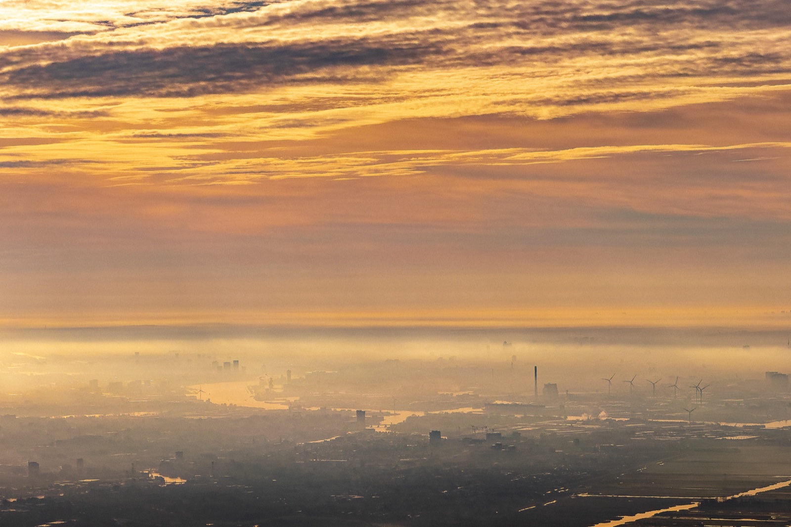 Vista aérea de una ciudad tendida bajo una capa de niebla al amanecer de otoño; el cielo, estratificado en nubes doradas, ocupa la mitad superior, y abajo un estuario devuelve la luz entre grúas, chimeneas y aerogeneradores.