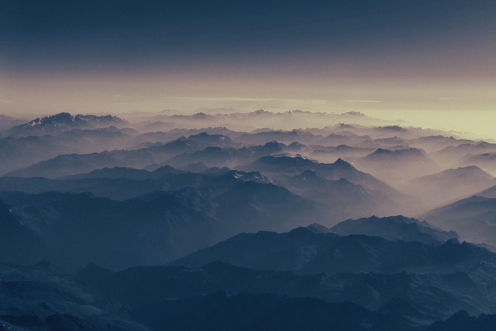 Vista aérea de una sucesión de cordilleras en tonos azules bajo la bruma del amanecer, con manchas de nieve en las cumbres más altas.