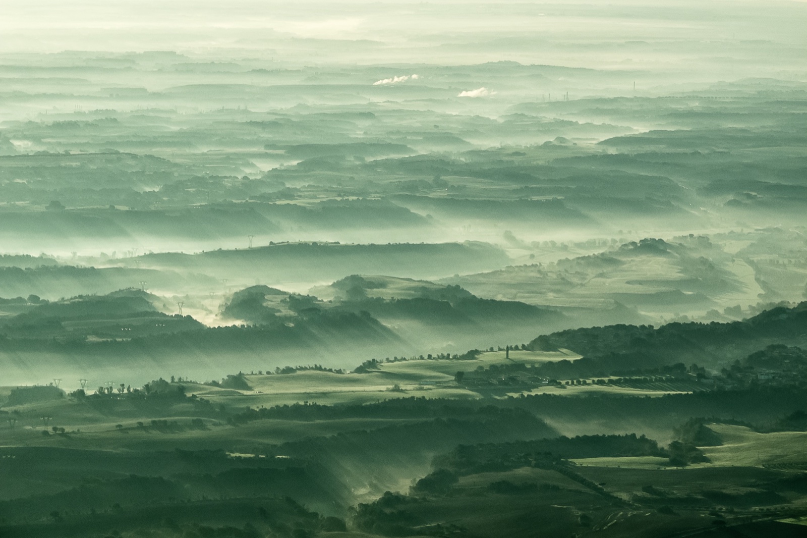 Vista aérea de una campiña ondulada en tonos verdes y azulados al amanecer; la luz rasante atraviesa la bruma y proyecta sombras largas que separan las lomas en capas sucesivas hasta el horizonte.