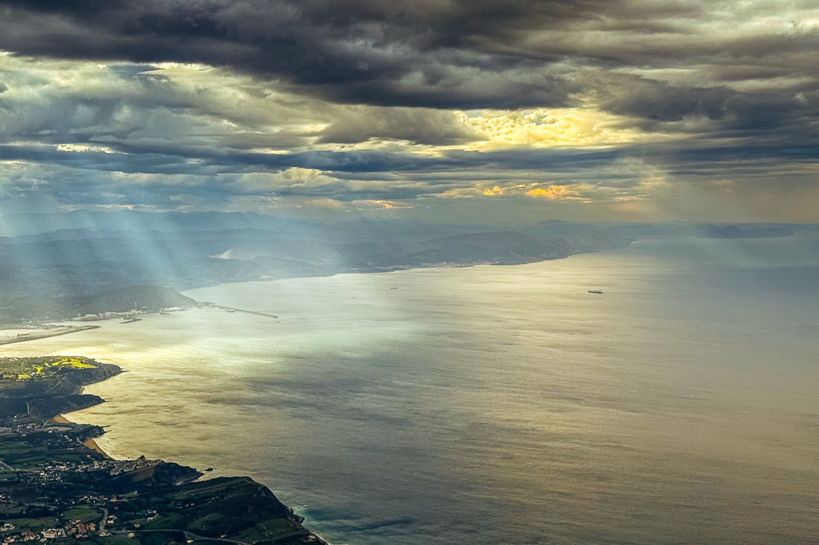 Vista aérea de una costa al ocaso con rayos crepusculares atravesando un techo de nubes oscuras; la luz cae en cortinas sobre el mar y dos barcos avanzan como puntos diminutos.